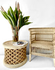 Natural Malawi Cane Chair beside carved round side table and green plant – Warm, welcoming corner styled with natural textures and layered aesthetics.